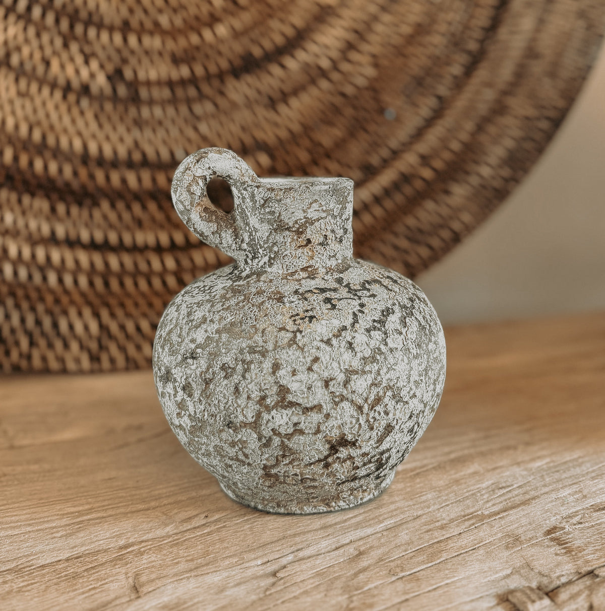 A small rustic terracotta jug vase with a rounded body, short neck, and loop handle, shown on a wooden surface with a woven rattan wall hanging in the background.