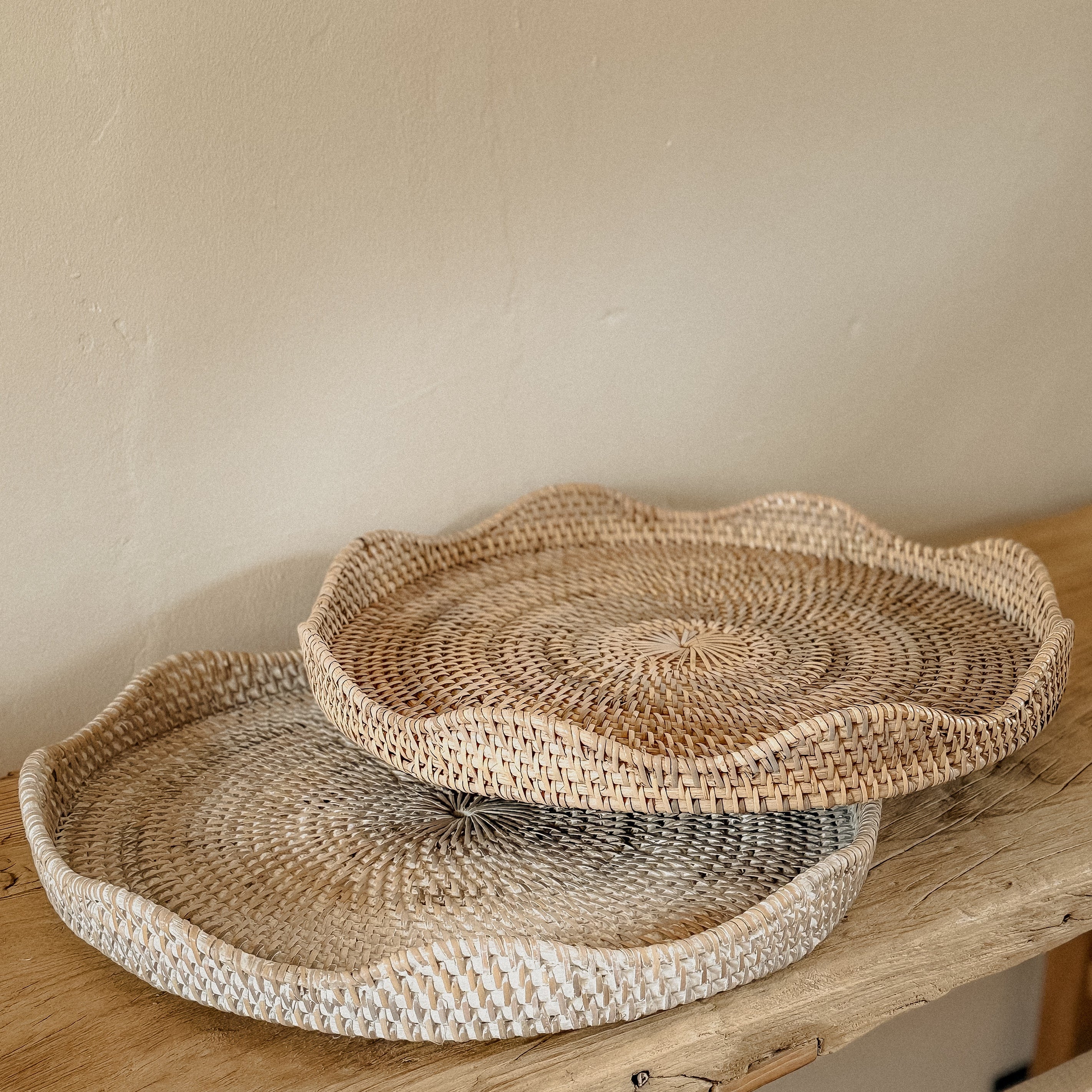 Whitewash round scallop rattan tray and natural round scallop rattan tray sat on a rustic elm console table with a light stone wall