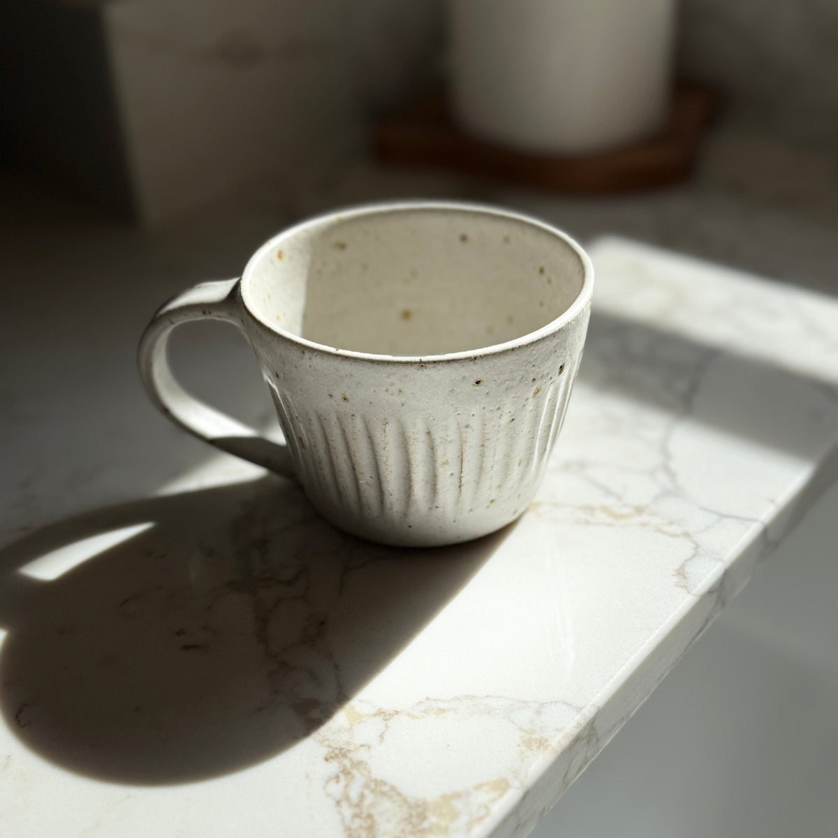 A speckled off-white ceramic mug with a ribbed exterior and a C-shaped handle, resting on a white marble countertop in direct sunlight.