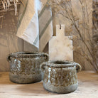 Two rustic handmade ceramic pots with side handles, featuring a textured earthy glaze, displayed on a wooden surface with a striped kitchen towel and dried botanicals in the background.