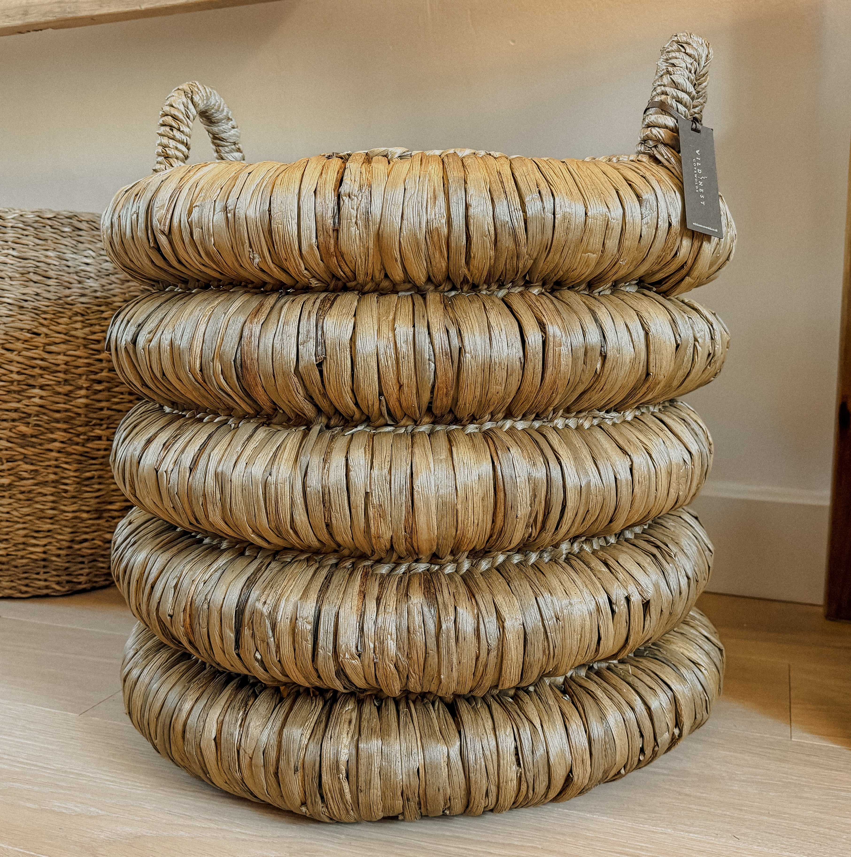 A chunky round woven basket made of water hyacinth with two loop handles, stacked on a light wood floor in an indoor setting.