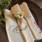Three cream-colored cotton napkins with printed illustrations and text, rolled up and tied with a small white flower on a woven brown placemat.
