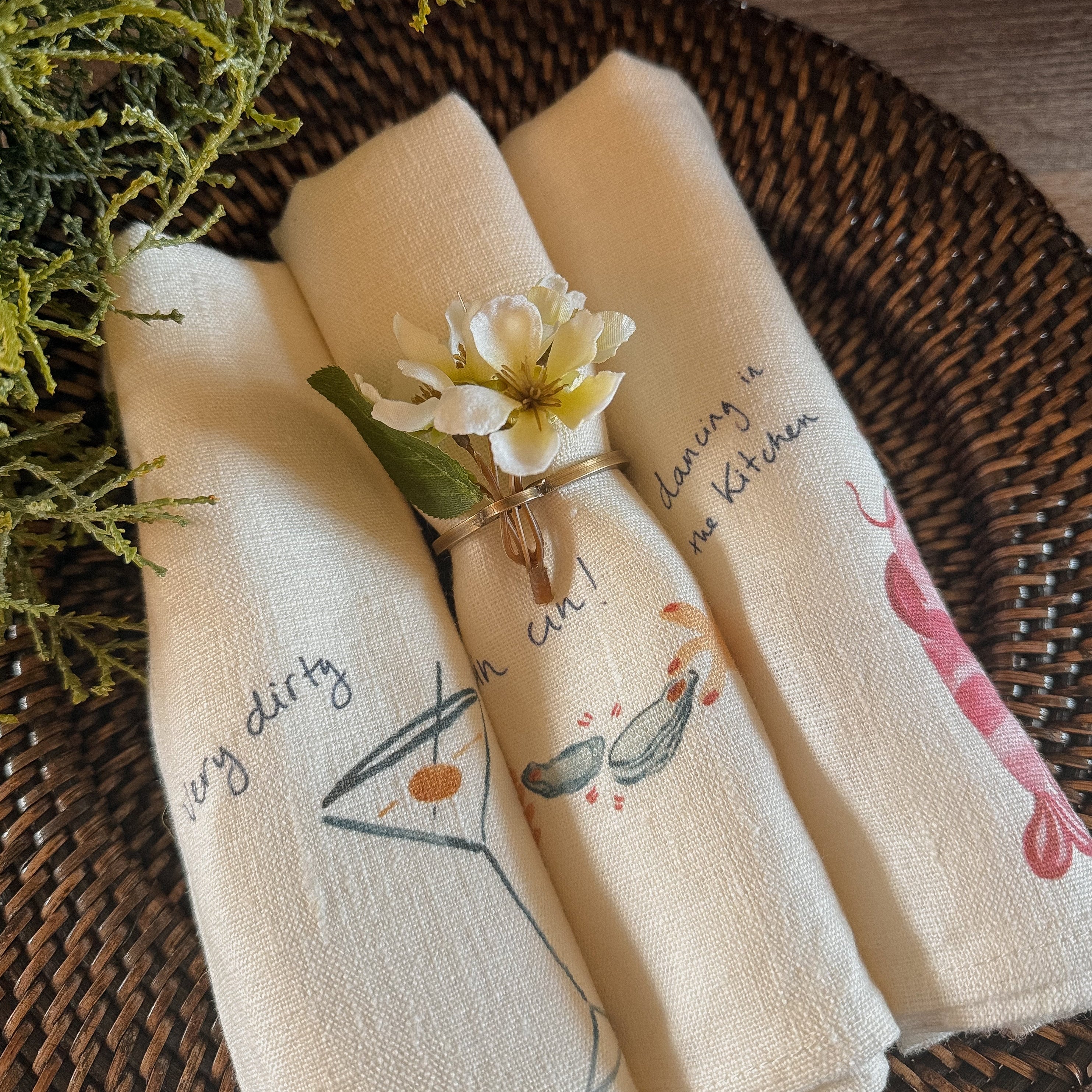 Three cream-colored cotton napkins with printed illustrations and text, rolled up and tied with a small white flower on a woven brown placemat.