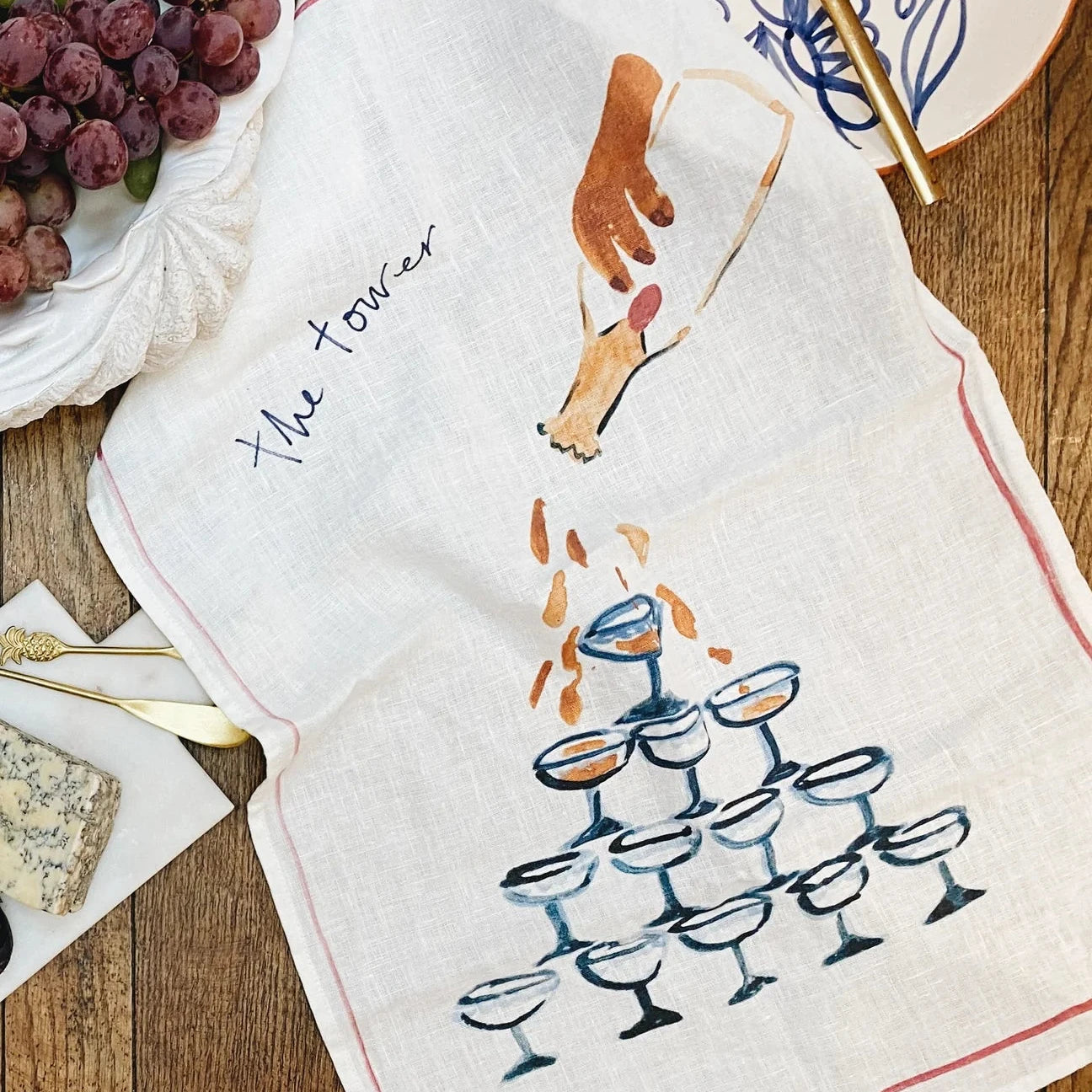 A decorative illustrated linen tea towel showing a tower of champagne glasses with champagne being poured from the top, ruffled on a wooden table top and surrounded by food items, cutlery and plates