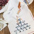 A decorative illustrated linen tea towel showing a tower of champagne glasses with champagne being poured from the top, ruffled on a wooden table top and surrounded by food items, cutlery and plates
