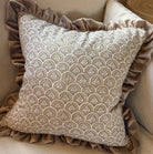 A square decorative throw pillow with a beige and taupe scallop pattern, featuring a ruffled taupe velvet border, styled on a beige fabric armchair.