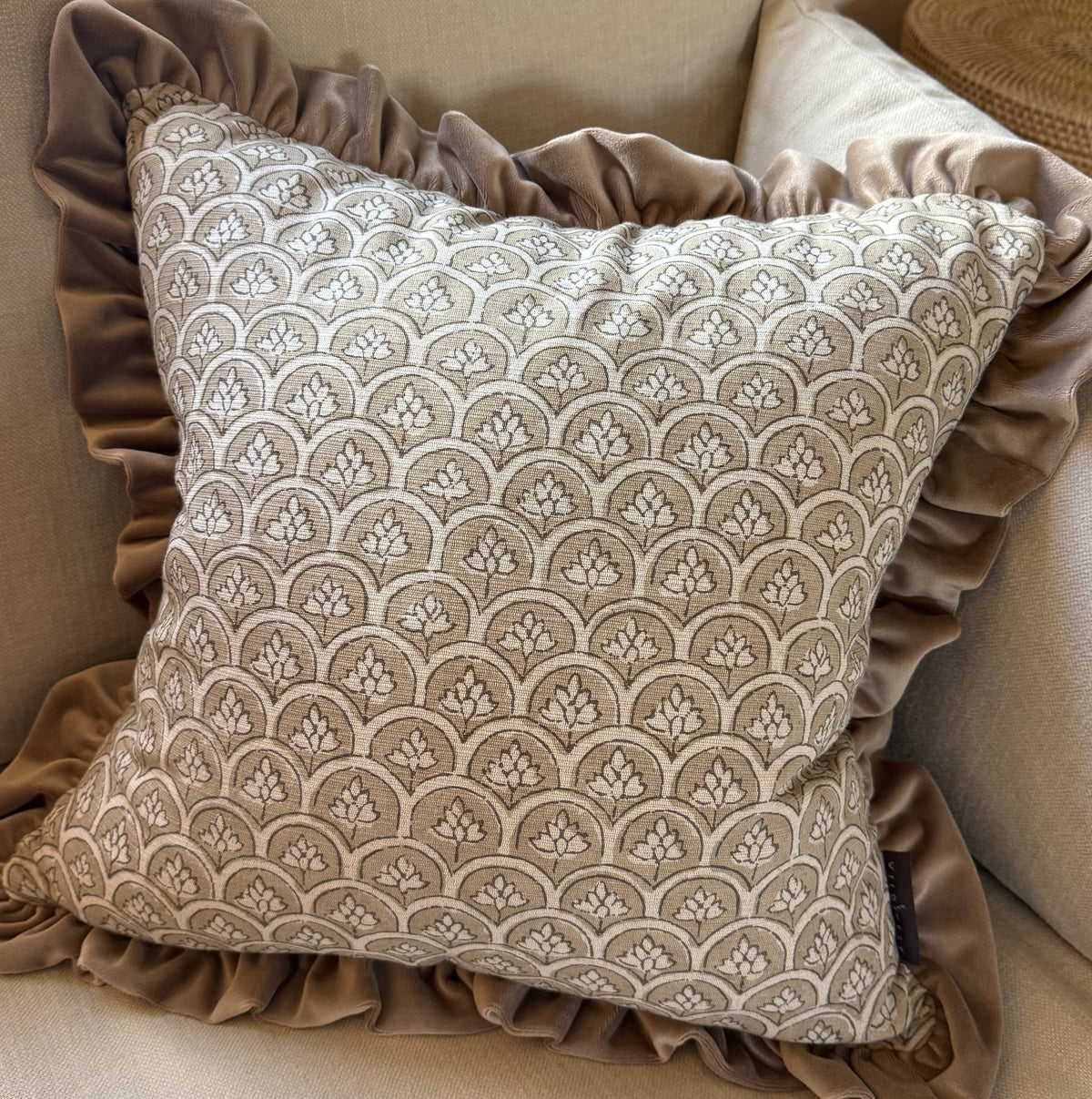 A square decorative throw pillow with a beige and taupe scallop pattern, featuring a ruffled taupe velvet border, styled on a beige fabric armchair.
