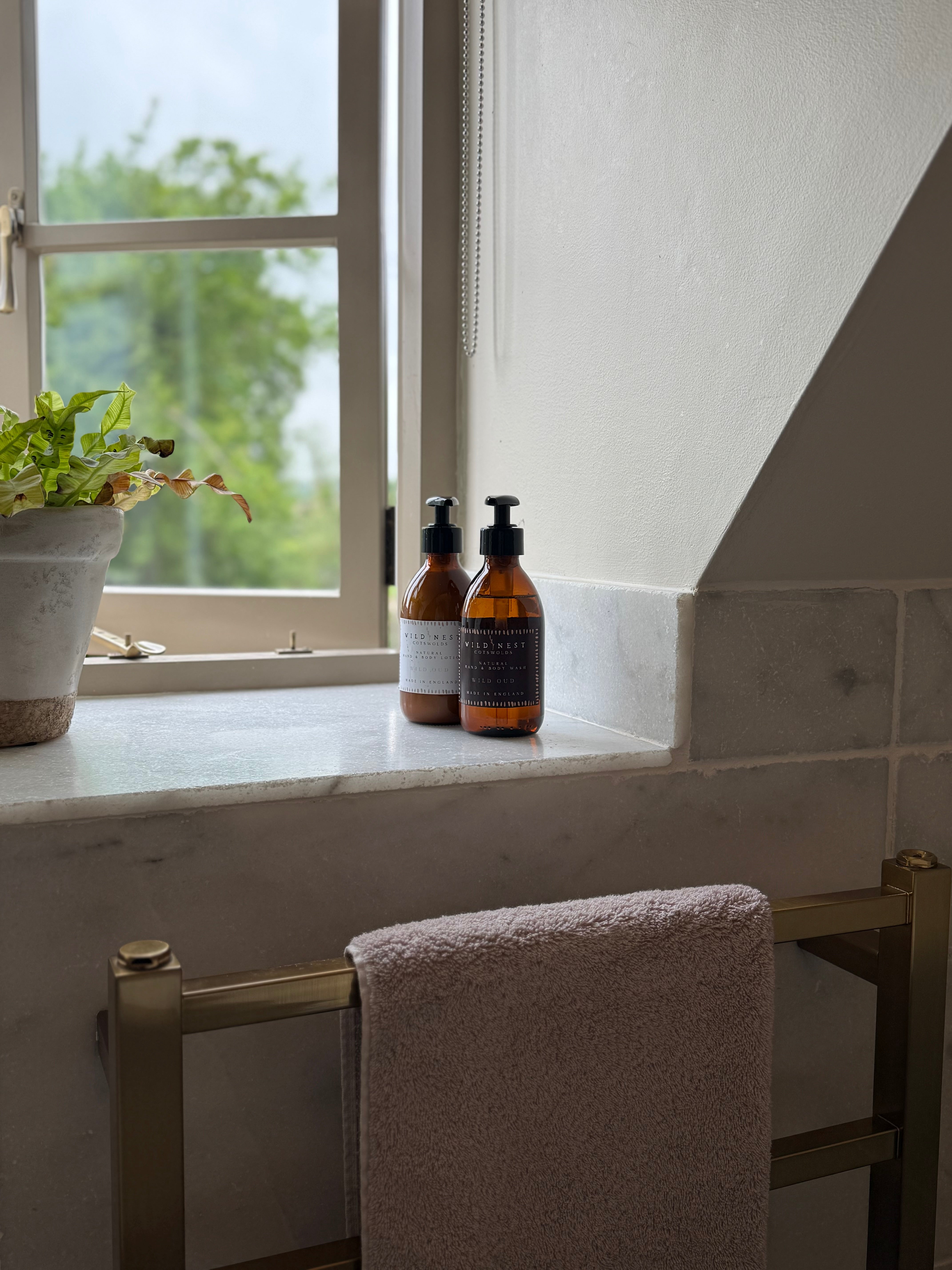 A Cotswold cottage bathroom scene with natural hand and body lotion bottles on a marble windowsill and a towel hanging on a brass radiator.