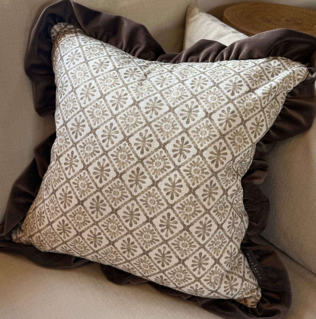 A square decorative throw pillow with a beige and brown geometric floral pattern, layered over a dark brown ruffled velvet throw, resting on a light beige sofa.