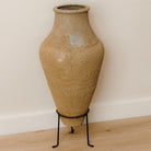 A large rustic vase reclaimed from Turkey with a rustic finish sat in a black metal stand on a wooden floor 