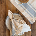 A styled flat-lay of a natural and denim striped linen napkin and matching napkins on a rustic wooden table, with a small white dish holding two round woven coasters.