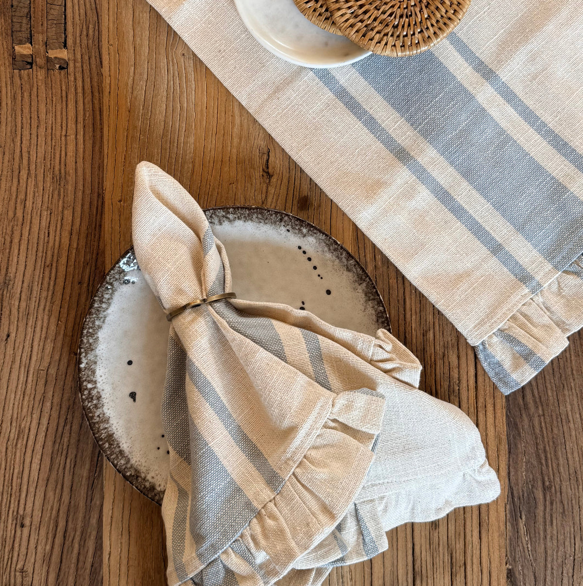 A styled flat-lay of a natural and denim striped linen napkin and matching napkins on a rustic wooden table, with a small white dish holding two round woven coasters.