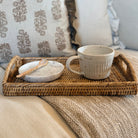 A rustic woven wicker tray holding a speckled ceramic mug and a matching small dish with a spoon, styled on a beige blanket with patterned throw pillows in the background.