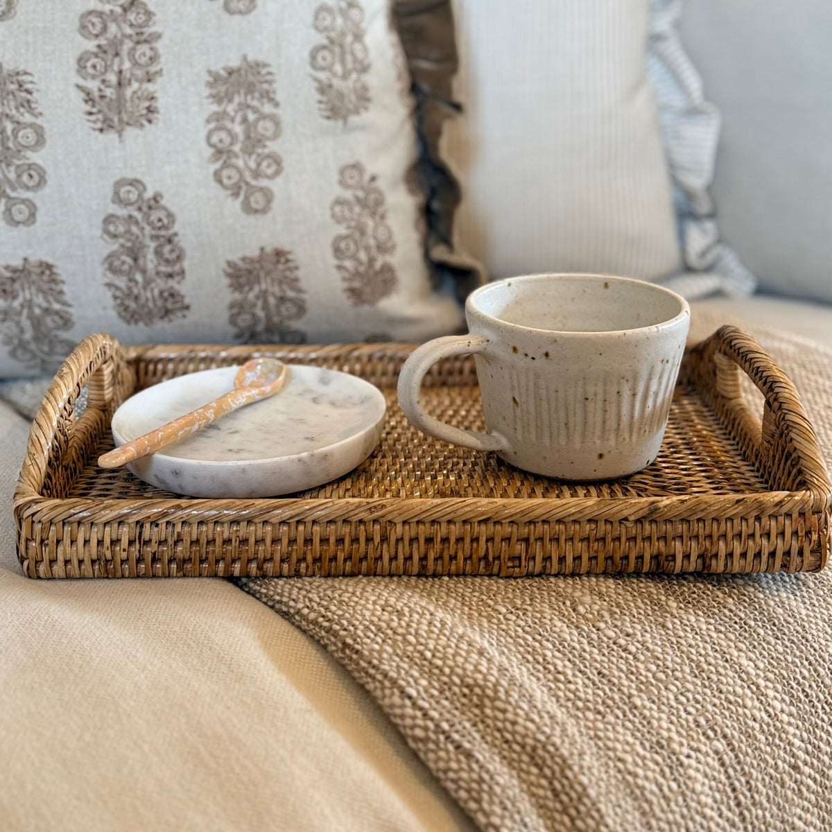 A rustic woven wicker tray holding a speckled ceramic mug and a matching small dish with a spoon, styled on a beige blanket with patterned throw pillows in the background.
