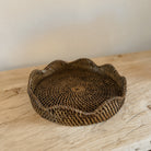 A round woven rattan tray with a scalloped edge, displayed on an elm wood console tabletop against a neutral beige wall.