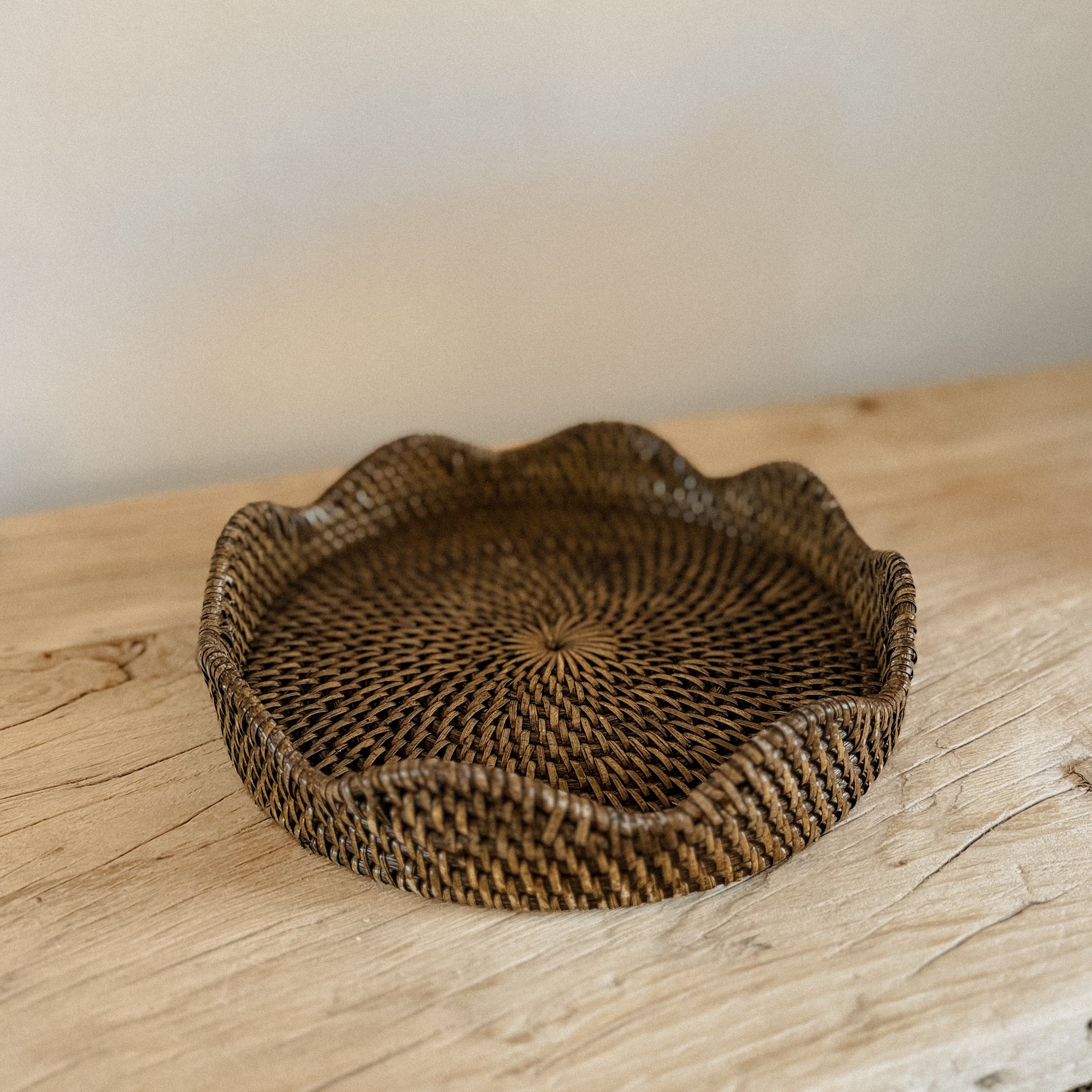 A round woven rattan tray with a scalloped edge, displayed on an elm wood console tabletop against a neutral beige wall.