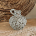 A small rustic terracotta jug vase with a rounded body, short neck, and loop handle, shown on a wooden surface with a woven rattan wall hanging in the background.