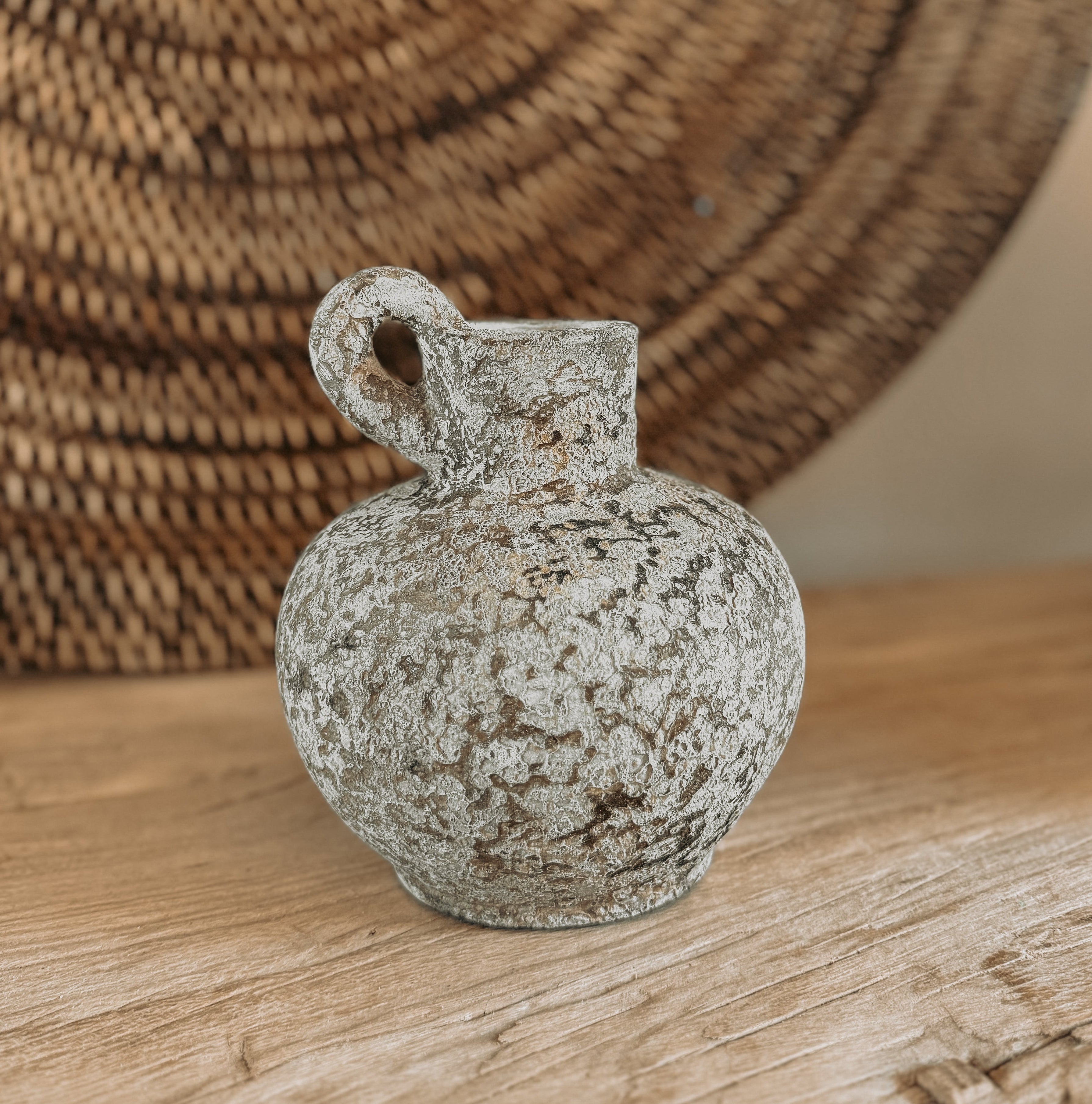 A small rustic terracotta jug vase with a rounded body, short neck, and loop handle, shown on a wooden surface with a woven rattan wall hanging in the background.