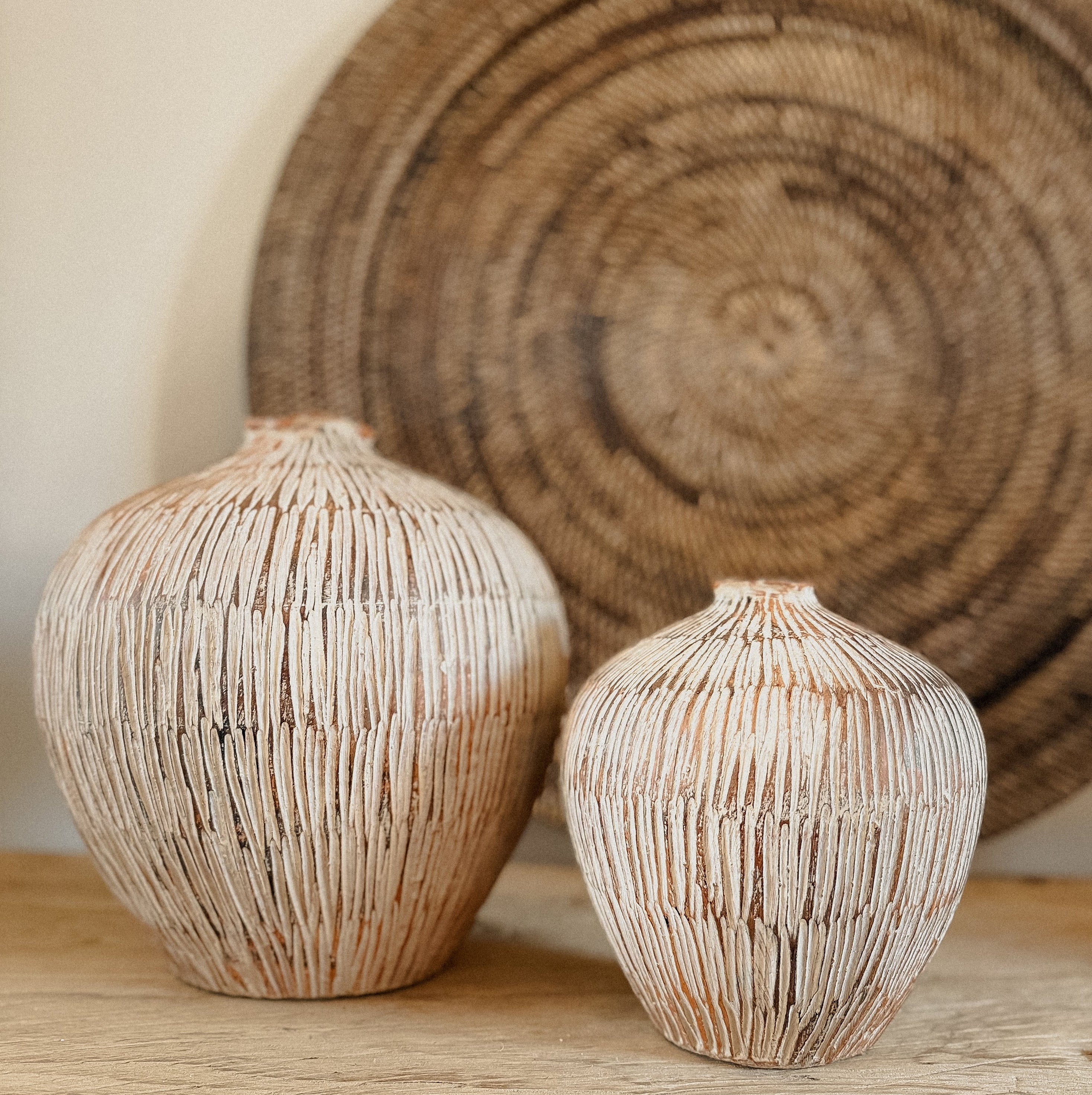 Two handcrafted terracotta vases with a carved design, one large and one small sat on an elm console table top with a dark brown circular rattan wall hanging behind. 
