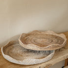 Whitewash round scallop rattan tray and natural round scallop rattan tray sat on a rustic elm console table with a light stone wall