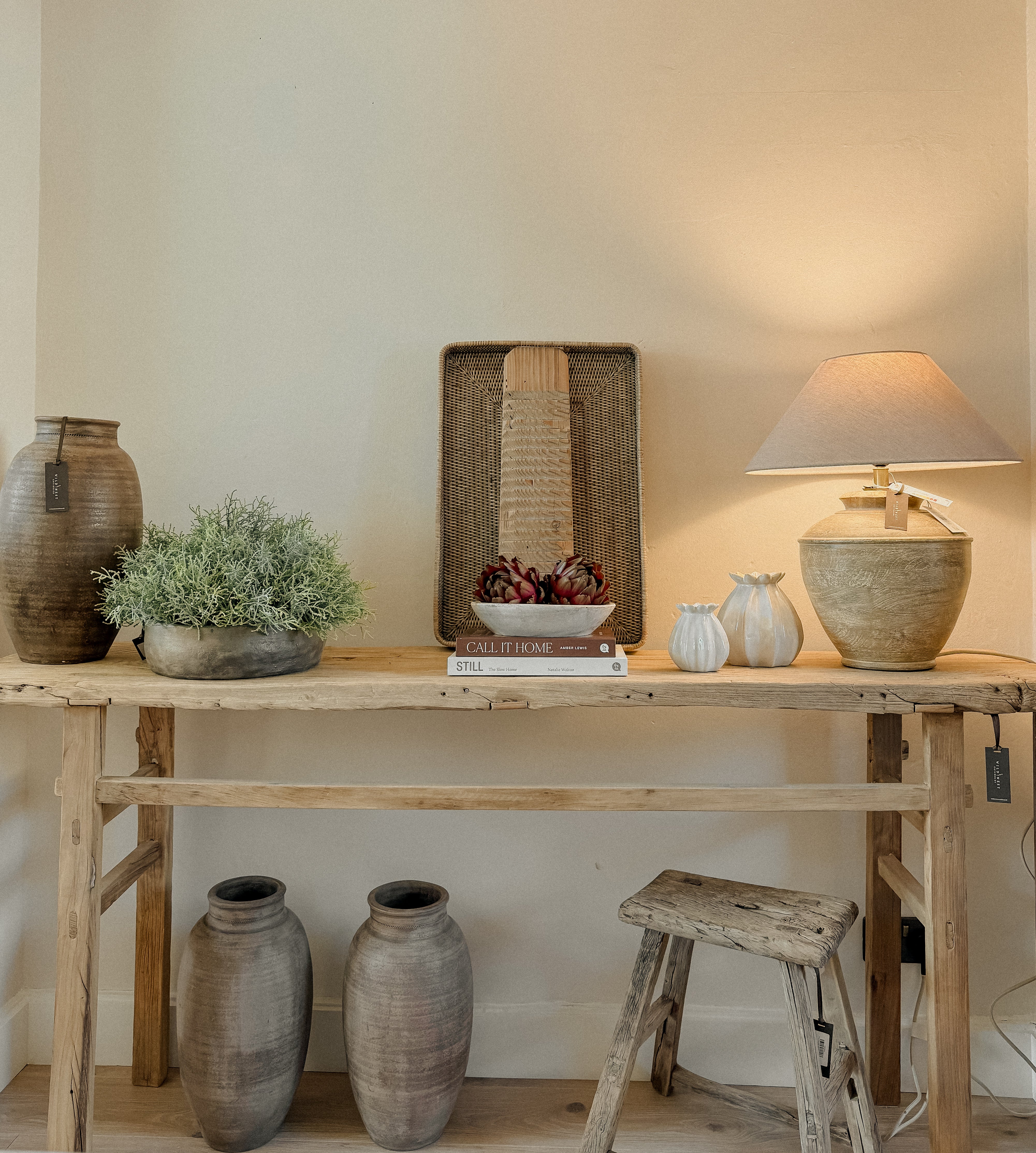 A rustic wooden elm console table styled with various home decor items including a turned wooden  table lamp, ceramic bud vases, a small potted green plant, and a woven rattan ottoman tray, set against a warm beige wall with a rustic elm stool below and two Turkish vases.
